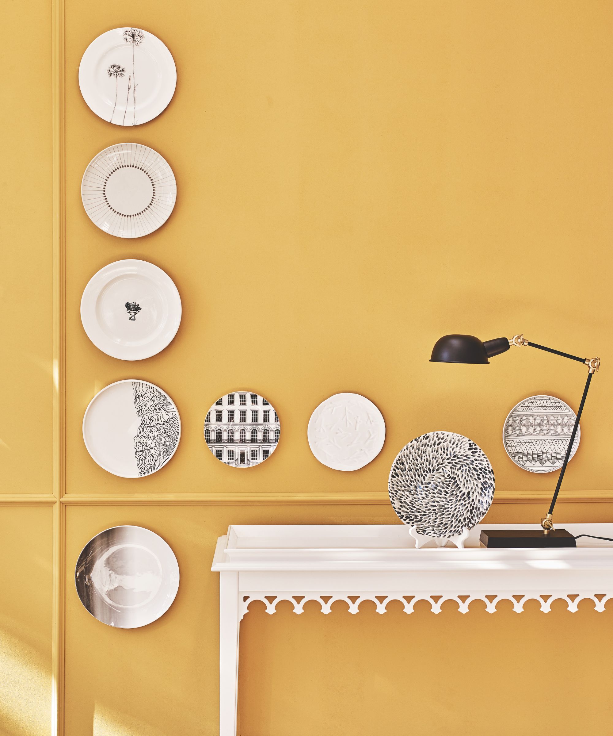 A yellow wall with decorative plates, and a white table in front of it with a black table lamp