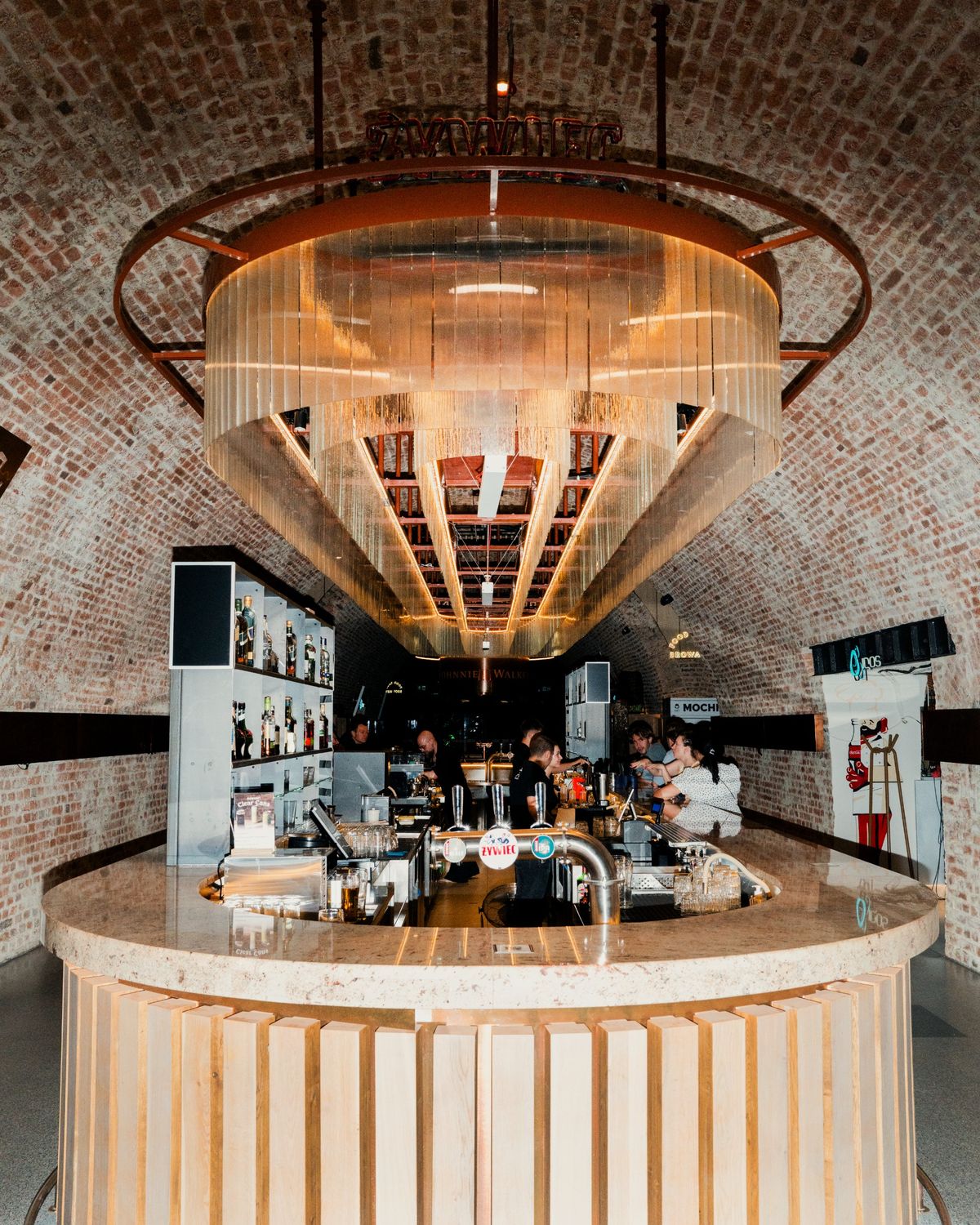 A symmetrical view of a curved wooden bar with &amp;#379;ywiec beer taps beneath a huge tiered glass chandelier, set inside a vaulted brick tunnel where drinker