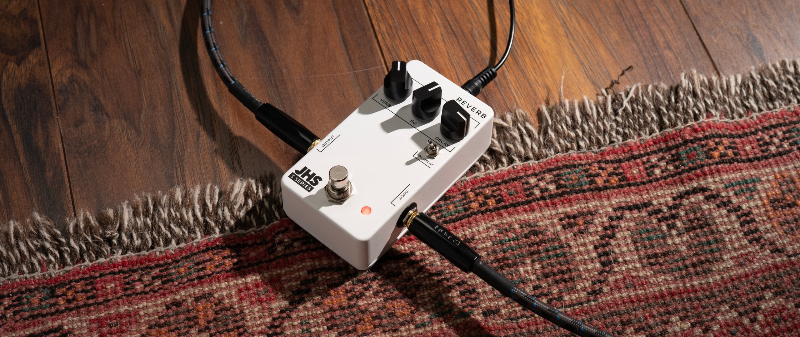 A JHS 3-Series Reverb pedal on a rug with guitar and power cables plugged in
