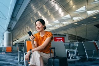 Woman with suitcase using smartphone while waiting for her flight at airport terminal.