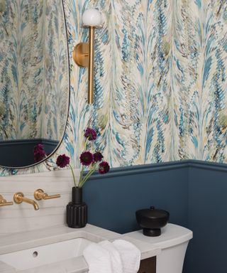 bathroom with blue wainscoting, blue patterned wallpaper above, brass fixtures, black accents, oval mirror