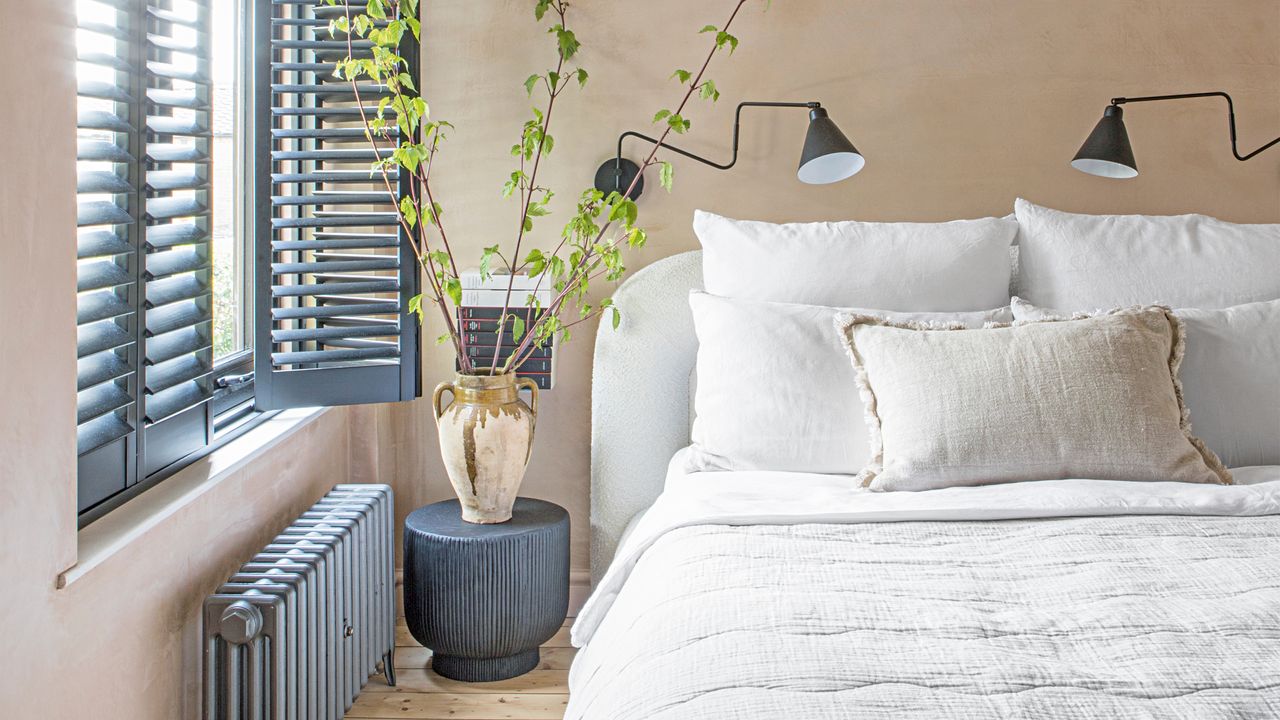 A bedroom with bare plaster walls and navy blue shutters on the windows with a bed dressed in white bedlinen