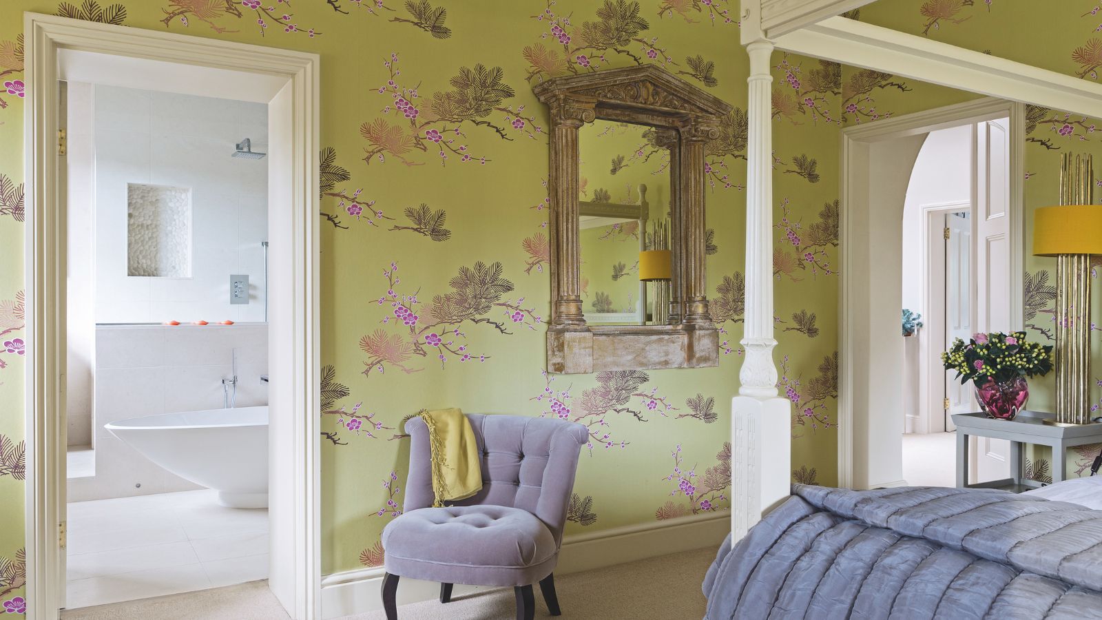 A green bedroom with statement wallpaper and a white four-poster bed with a grey blanket. To the left is a doorway open to a white en-suite bathroom, and a grey upholstered chair.