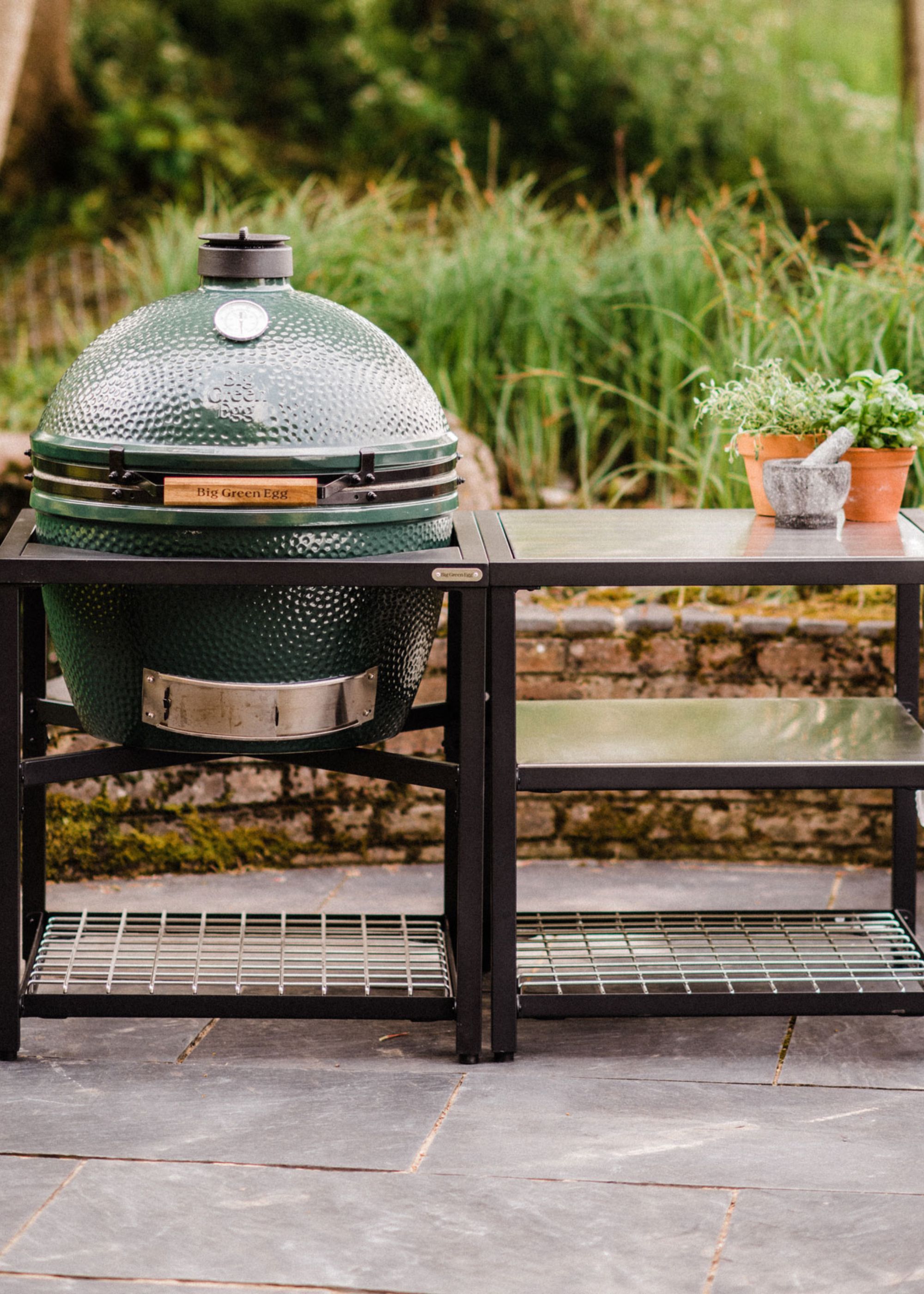 A big green egg inside a black grilling table beside two herb pots in a garden
