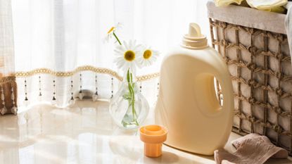 A bottle of plain yellow laundry detergent next to a wicker laundry hamper, a cloth, and a small vase of large dasies
