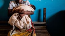a woman sits on a bench holding a baby wrapped in a blanket, whose face is turned from the camera