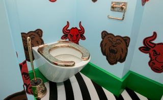 Always Close installation by Studio Job in Luxembourg. A bathroom with lion and bull heads printed on a blue wall, a toilet with a gold seat and a gold toilet cleaner.