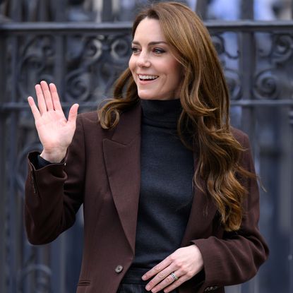 Kate Middleton wearing a brown blazer and black turtleneck waving and smiling in front of a metal gate