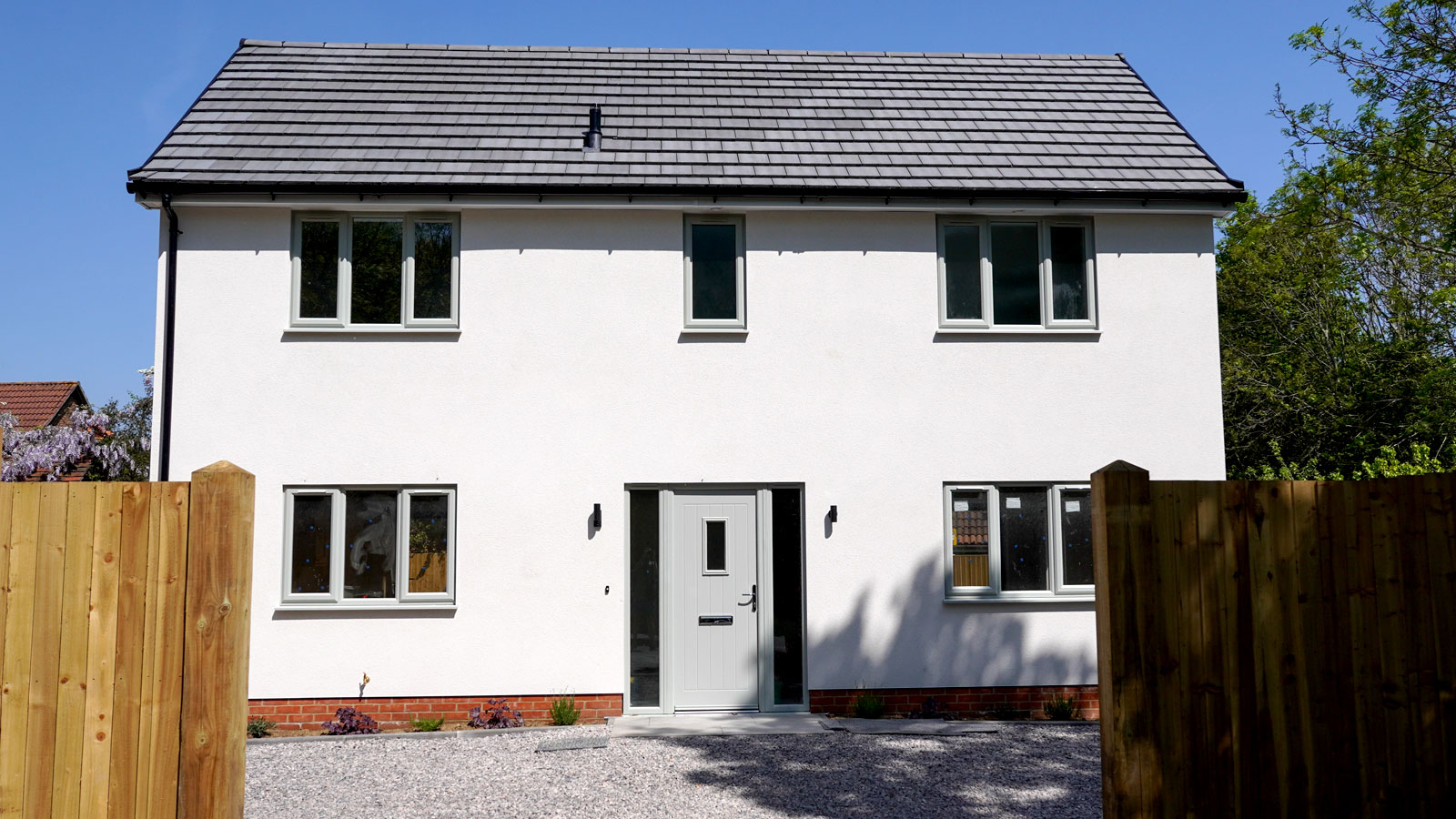 New two-storey house with white render and grey windows and doors and gravel drive with wooden fence in foreground