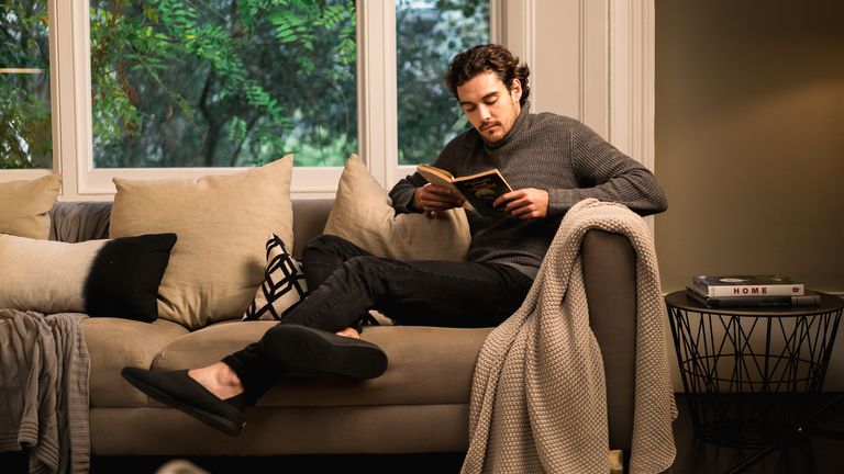 A man, relaxing on the sofa reading a book wearing black slippers – a paid of the best slippers for men you can buy