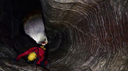 A person wearing a yellow helmet and red jacket repels down a dark cave.