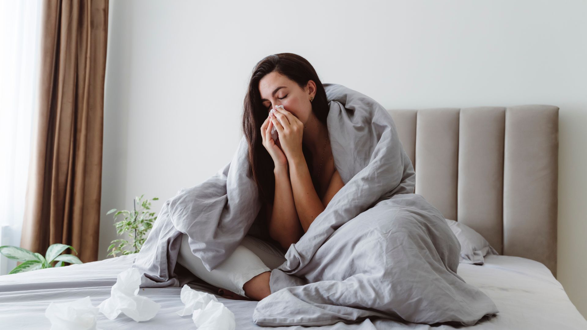 A woman sitting in bed wrapped up in her duvet as she blows her nose.