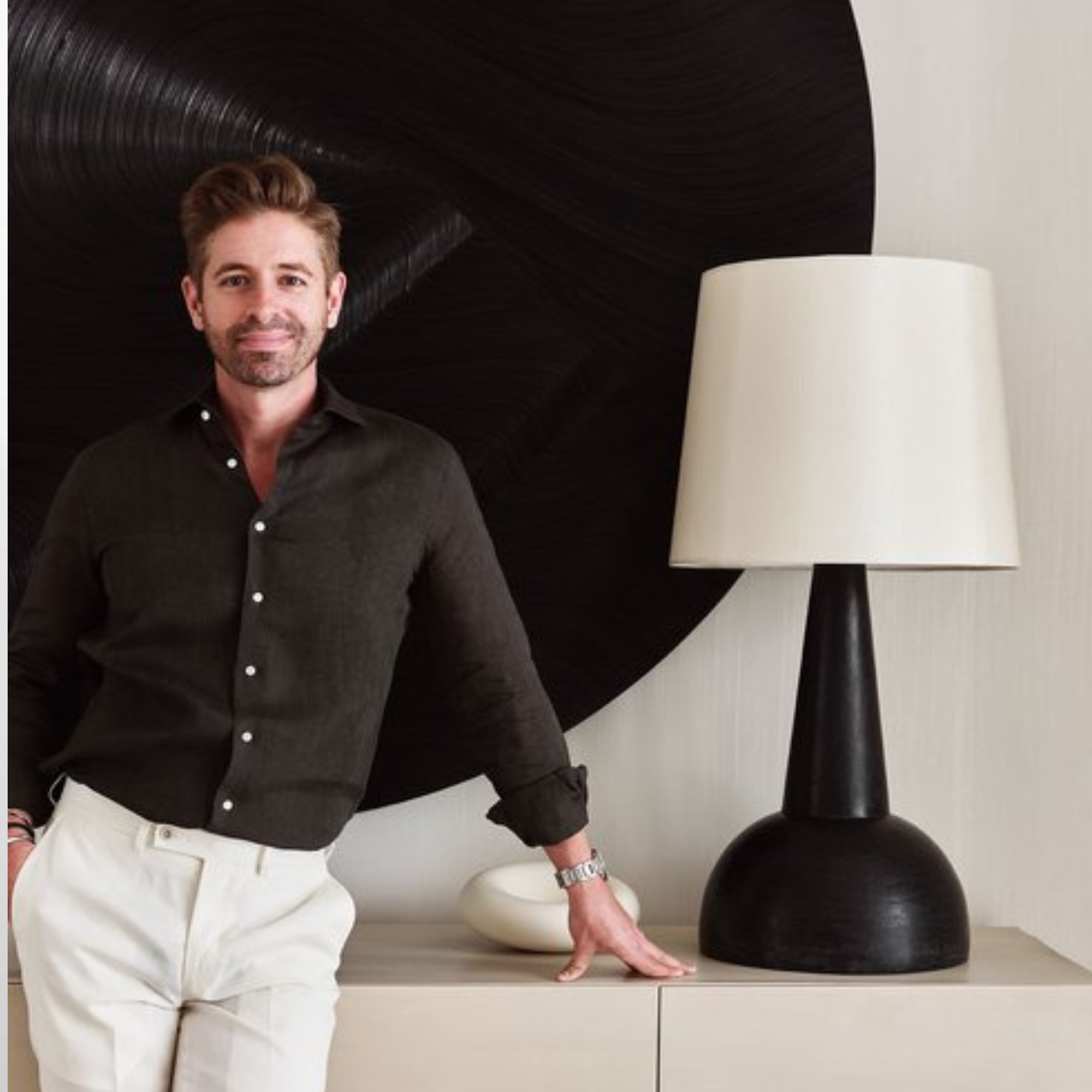 A photo of designer Michael Ellison leaning against a table next to a large black-and-white lamp.