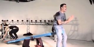 Steve-O dressed in blue jeans and a grey shirt getting hit in the nuts with a makeshift seesaw after someone jumped on the other end.