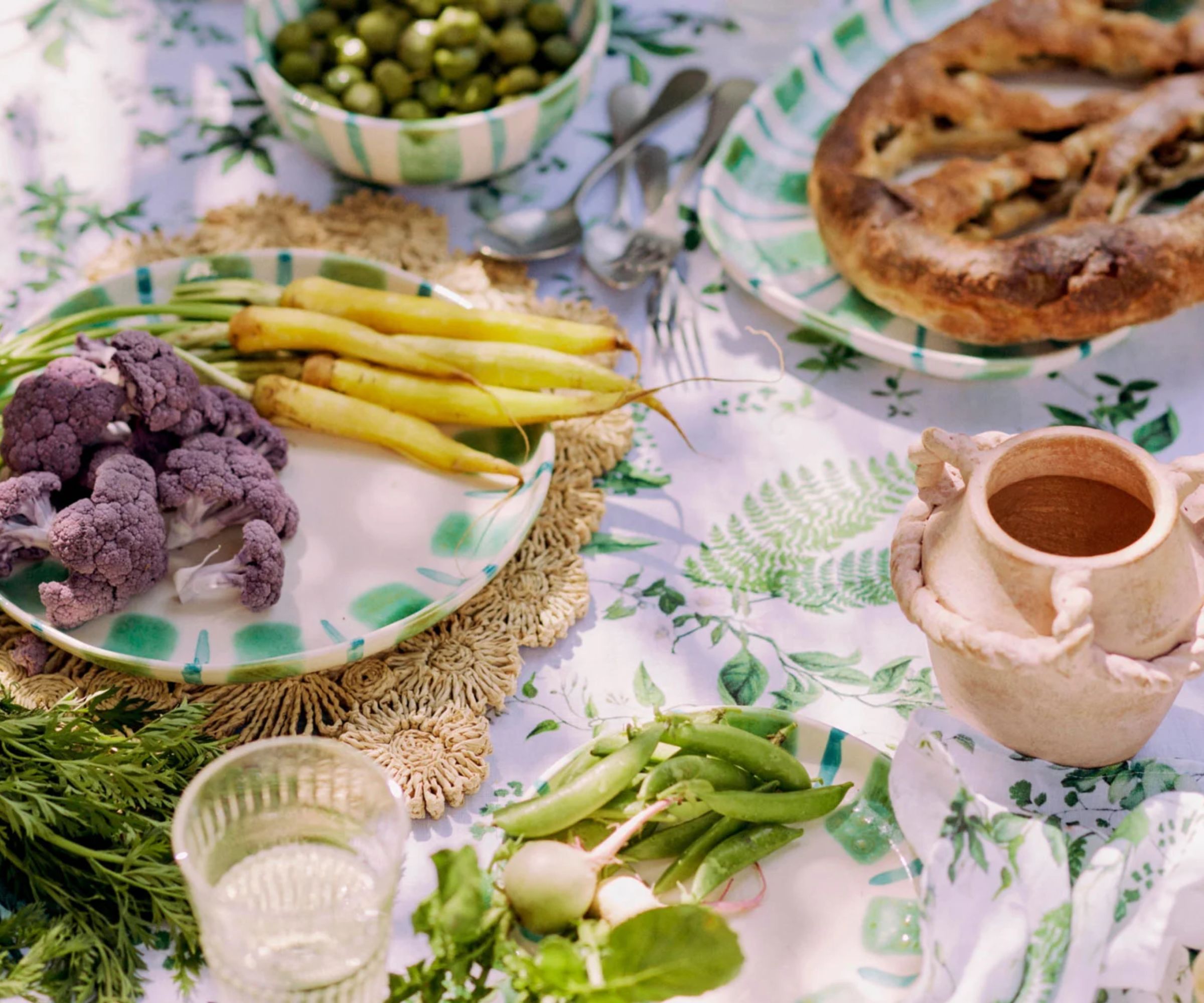 Mediterranean style tablescape with foliage print table cloth, rattan placemat, terracotta vase, and green painted platters with fresh produce