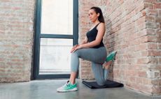 Woman half kneels next to an exposed brick wall, stretching her hips. She wears a black tank top, grey sports leggings and trainers. Her rear knee rests on a folded up black yoga mat.