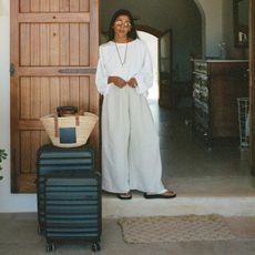Woman in white two-piece set standing in front of doorway with Loewe raffia bag and two pieces of black carry-on luggage.