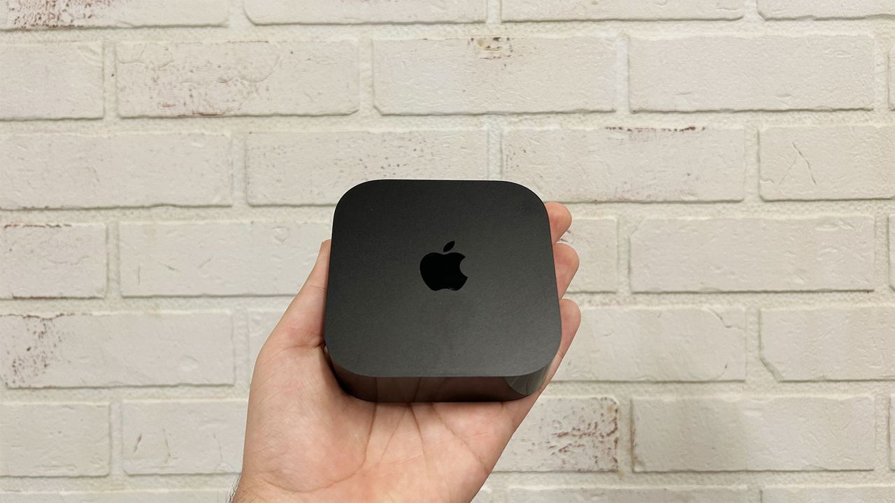 A hand holding a black Apple TV 4K streamer in front of a white brick wall.