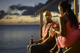 DI Mervin Wilson (Don Gilet) sits on the steps leading up to Catherine's Bar from the beach at twilight with DS Naomi Thomas (Shantol Jackson). Naomi is holding a piece of paper in her hands, and Mervin is clutching a bottle of beer.