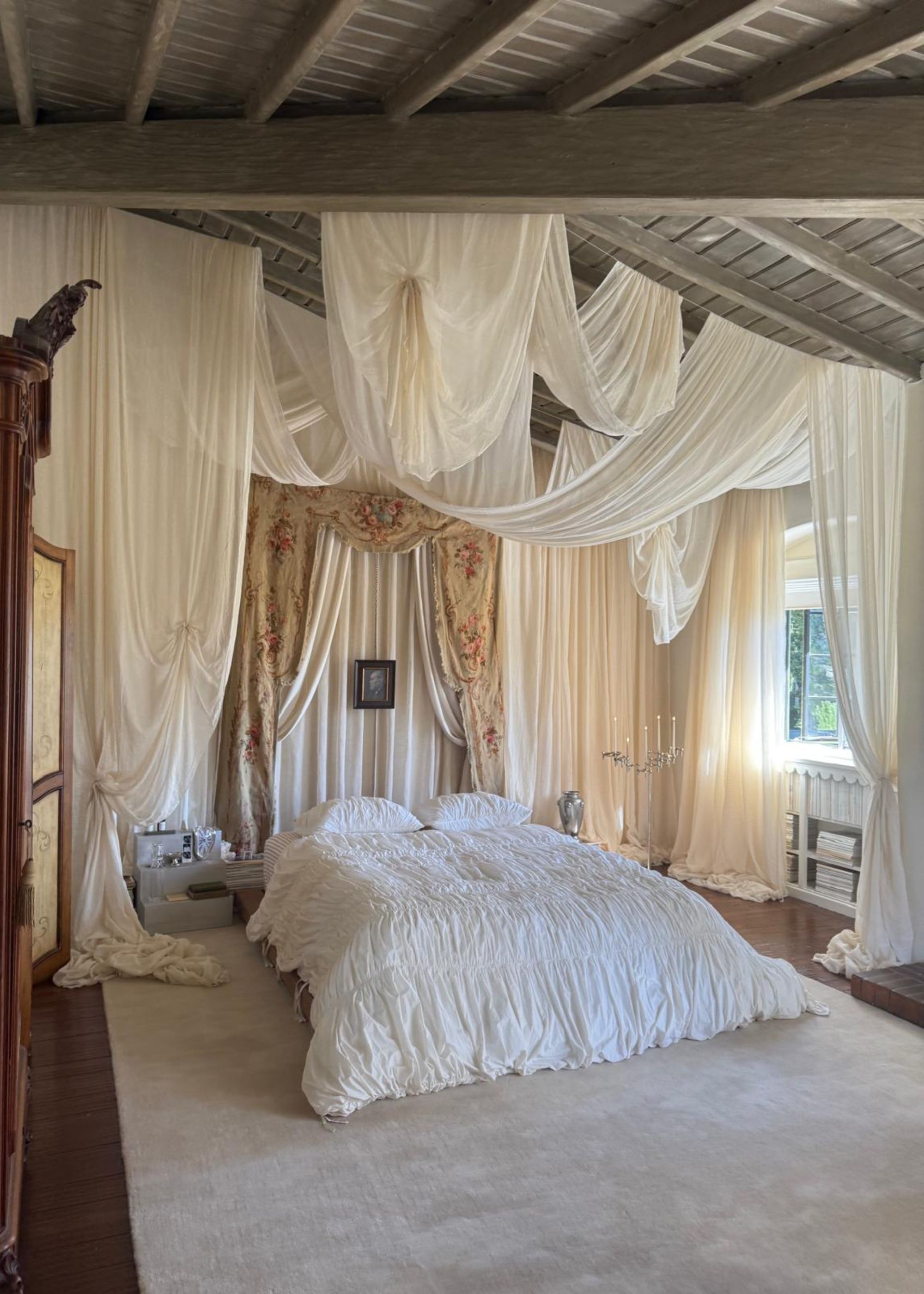 A large neutral bedroom with a sloped wood ceiling. A large cream rug on the floor and a large bed with white bedding. Attached to the ceiling, there is a canopy made from flowy, cream cotton draped between poles.