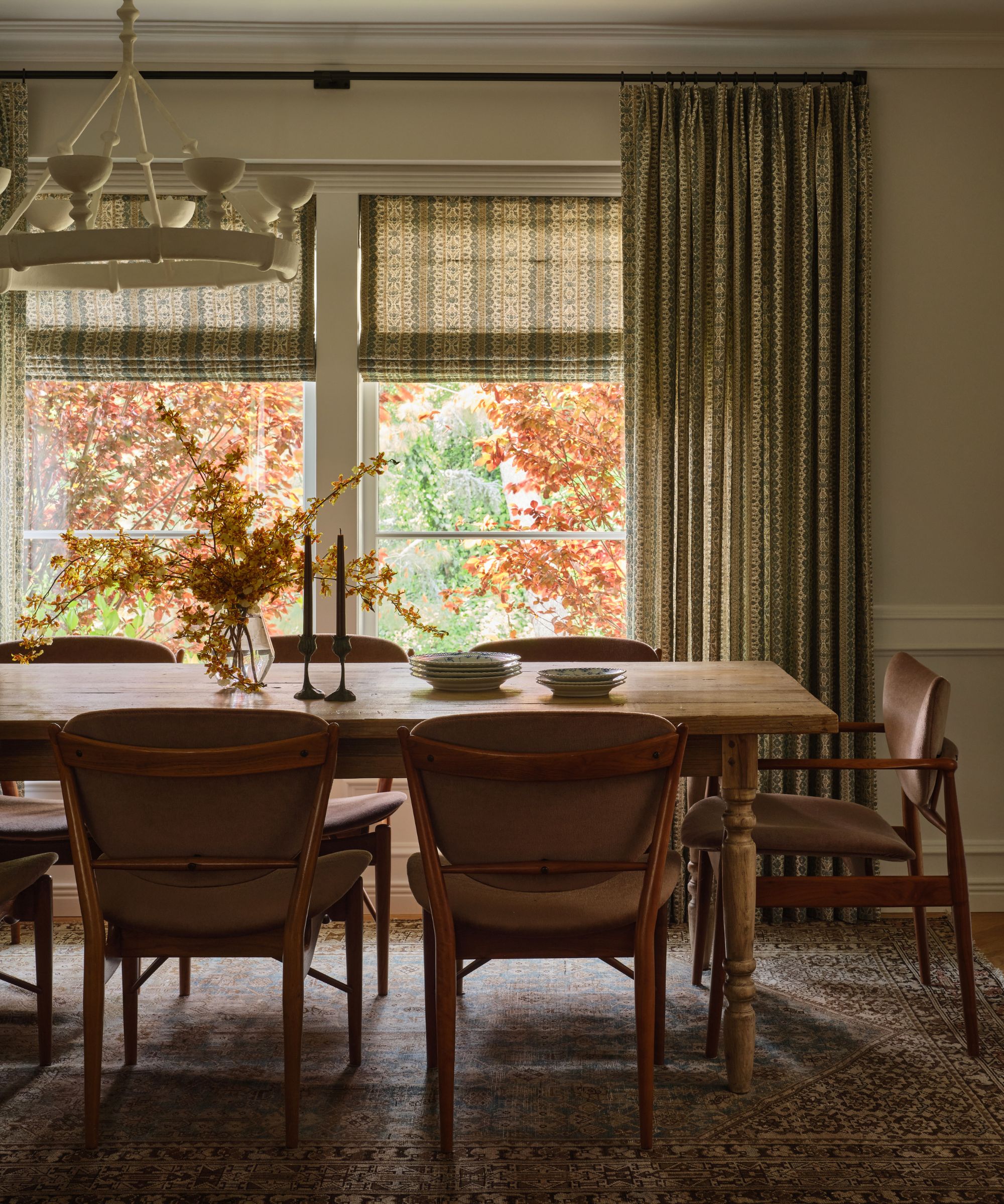 Dining room with a wooden table set for dinner, mid-century style chairs, a white chandelier, and windows with roman shades and curtains overlooking red and orange autumn foliage