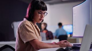 Female software developer using AI coding tools on a desktop computer with colleagues in background in an open plan office space.