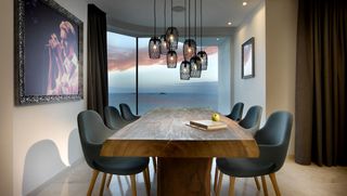 Natural solid wood long dining table , six dark grey chairs,, book and apple on the table top, framed picture of David Bowie on the left wall, black ceiling lights suspended over the table, brown curtains, window with view of the sea and red and blue sky, ceiling spotlights