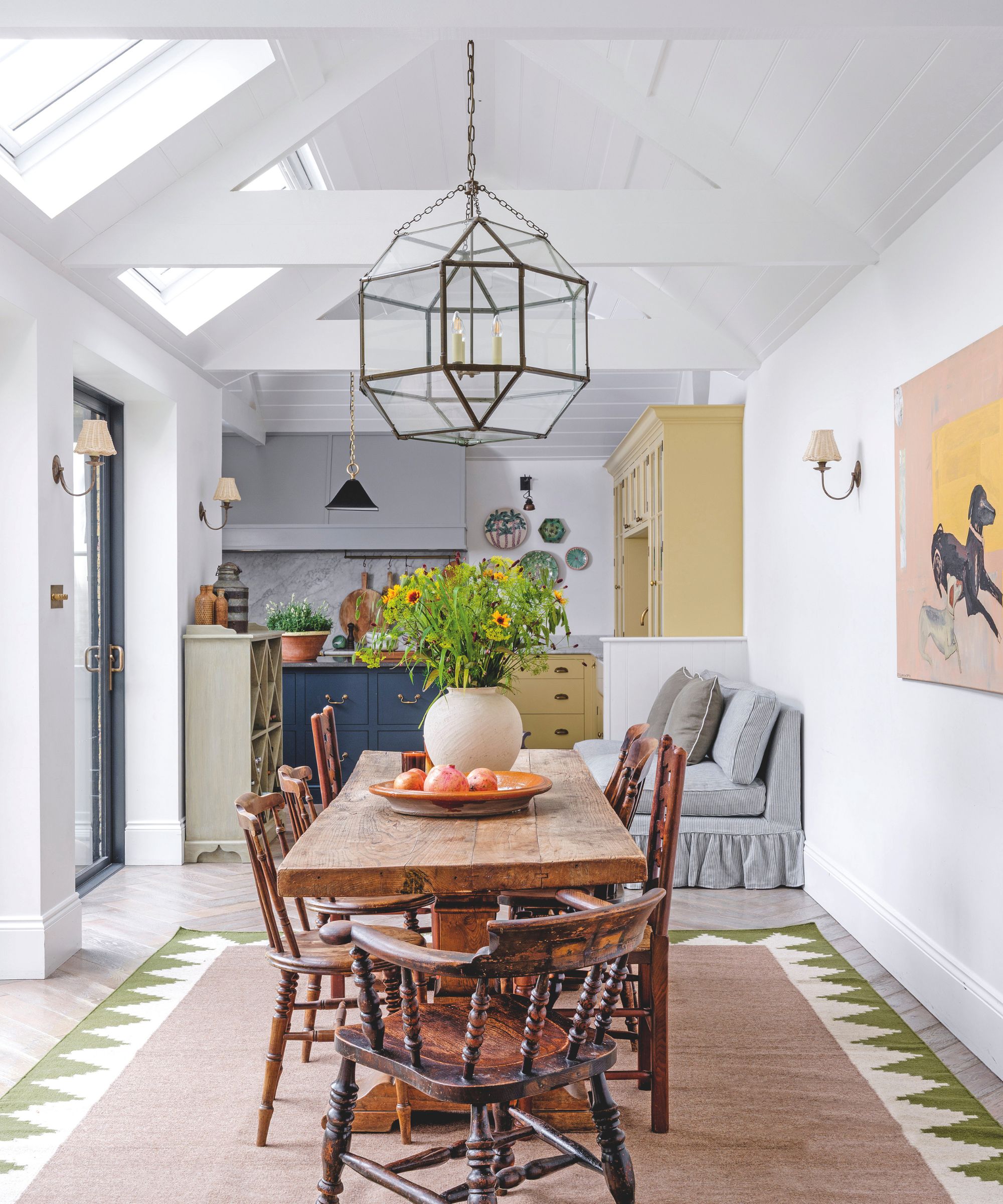 A bright, airy dining space with a rustic wooden table, antique spindle chairs, and a large glass lantern pendant light hanging from a vaulted white ceiling.