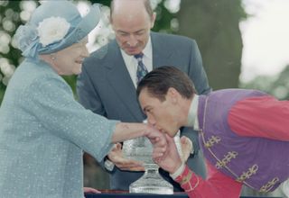 The Queen Mother wearing a blue coat and hat holding her hand out for a jockey to kiss it in 1997