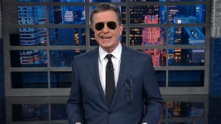 Stephen Colbert dons sunglasses in side for his late night monologue on April 23rd. He has on a suit and tie.