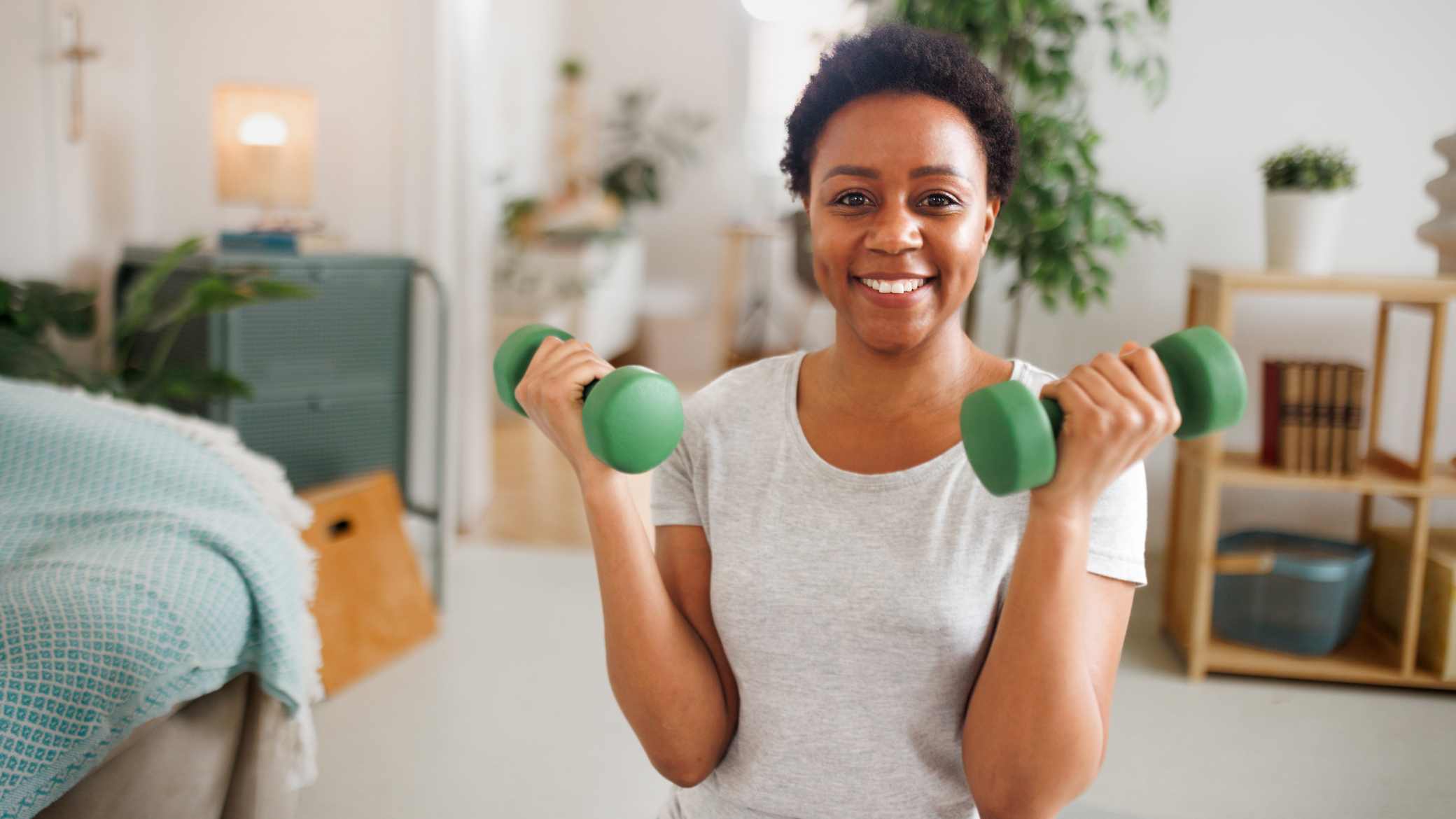 A woman kneels in a bedroom holding light dumbbells at her shoulders and smiling. 