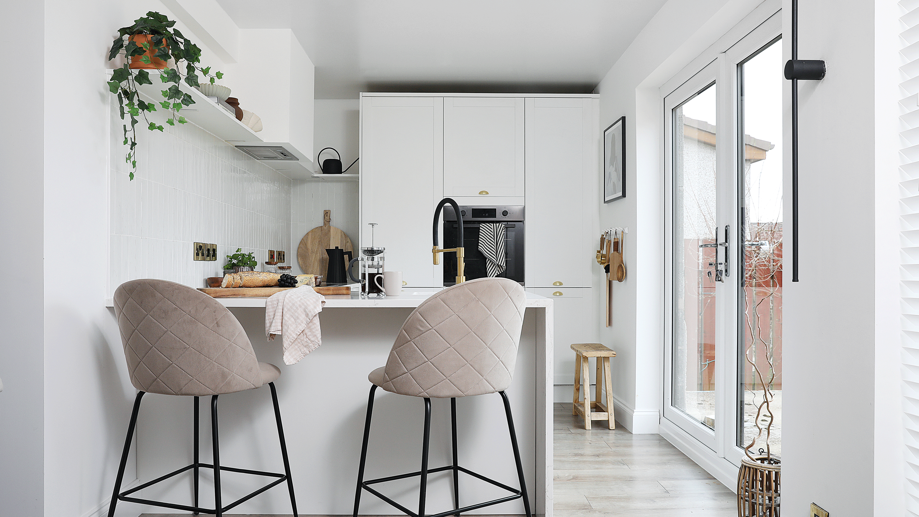 a small white shaker-style kitchen with pale grey wooden flooring and a peninsula breakfast bar with beige quilted upholstered bar stools