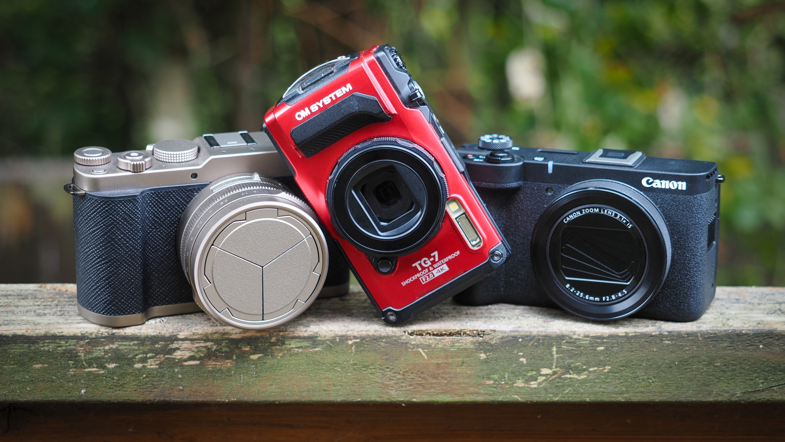 Panasonic Lumix L10, OM System Tough TG-7 and Canon PowerShot V1 compact cameras on a wooden surface outdoors