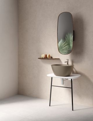 Room with neutral walls and white floor, beige wash basin on a white table top with two black metal legs, silver tap. long oval shape wall mirror reflecting fanned plant leaves, small wooden shelf with circular shaped soaps
