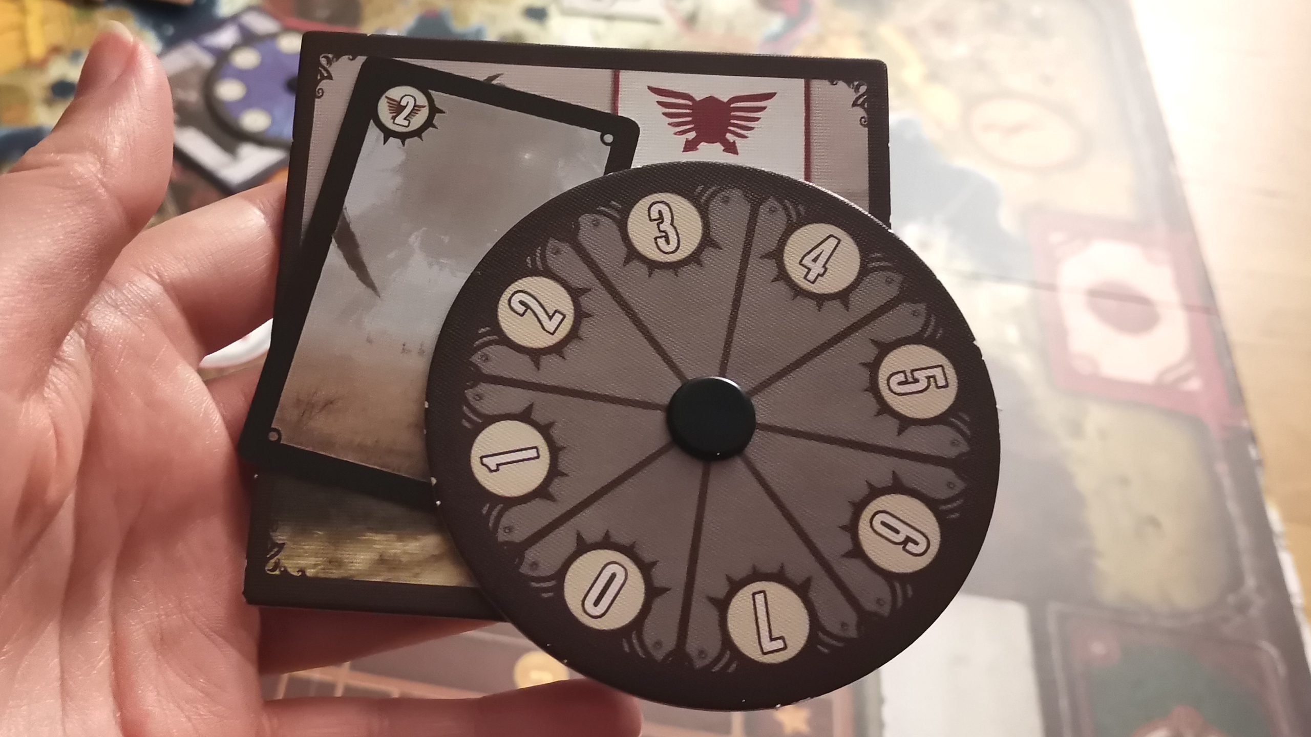 A hand holds a number spinner and cards from Scythe, with the board visible in the background
