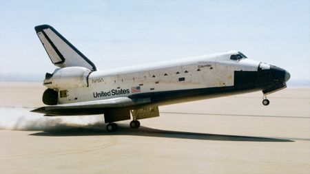 a black and white winged spacecraft touches down on its main gear on a dry lakebed