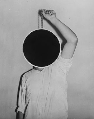 Black and white photo of man holding black circle by hanging chain which covers his face.