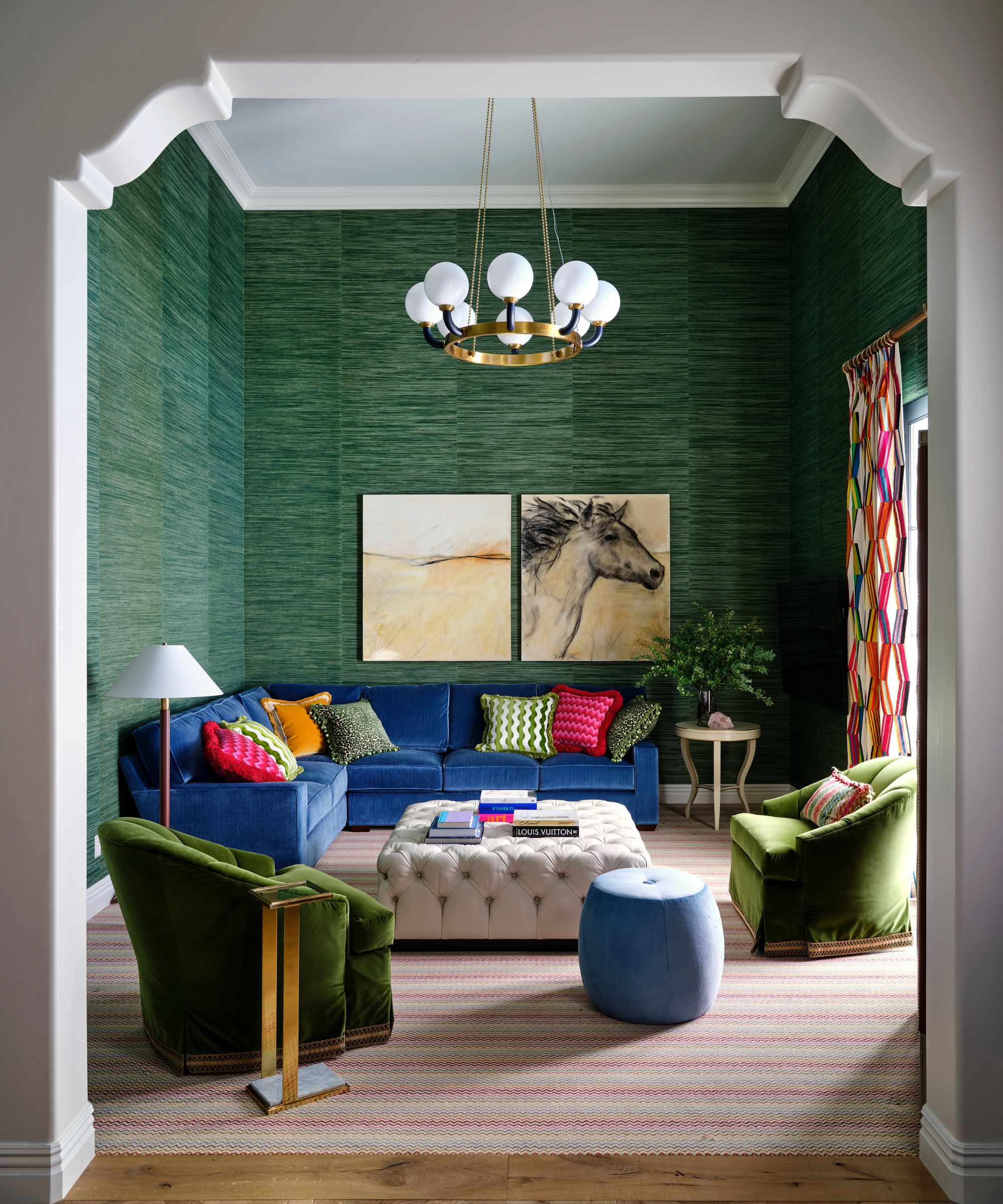 Living room with green grasscloth walls, velvet blue sofa, velvet green armchairs and quilted ottoman, with colourful pink and orange curtains and throw cushions