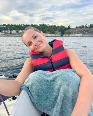 Princess Ingrid Alexandra sitting in a boat wearing a red and blue lifevest with her legs covered with a blue towel and the water and trees behind her