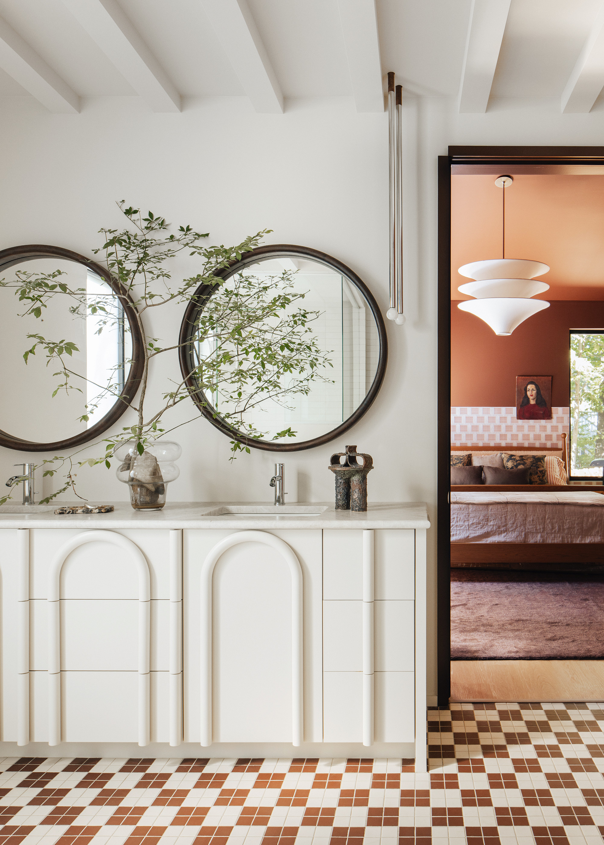 View from a white bathroom with carved vanity units and twin round mirrors into a terracotta bedroom.