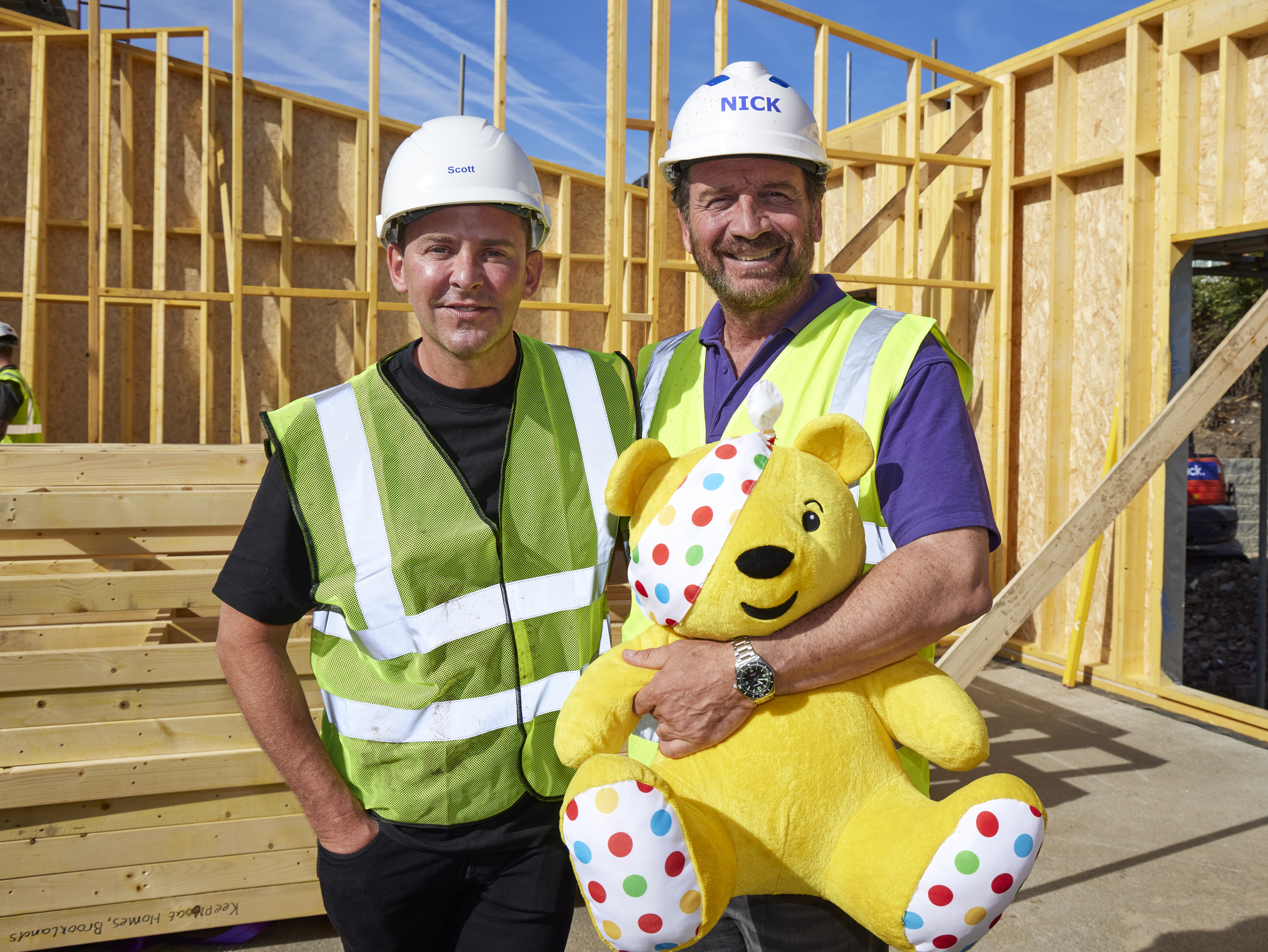 Scott Mills and Nick Knowles stand on the building site, wearing high-vis jackets and hard hats.  Nick is holding on to a stuffed Pudsey Bear