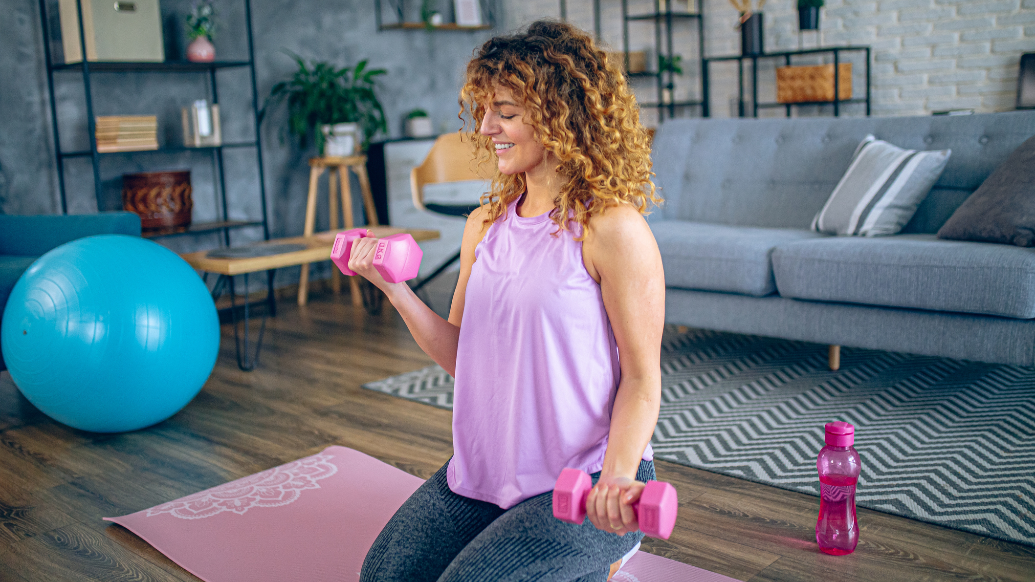 Woman kneels on an exercise mat in a living room holding small pink dumbbbells