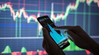 person looking at stock market chart on smartphone and large PC screen