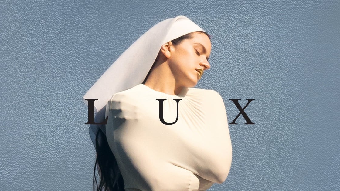 A person wearing a white, nun-like habit and gold lipstick poses with their eyes closed against a textured blue background, overlaid with the large black letters "LUX."
