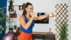 woman in a living room setting wearing a bright blue top and red bottoms and holding a small dumbbell in each hand and punching forward with one hand while smiling.