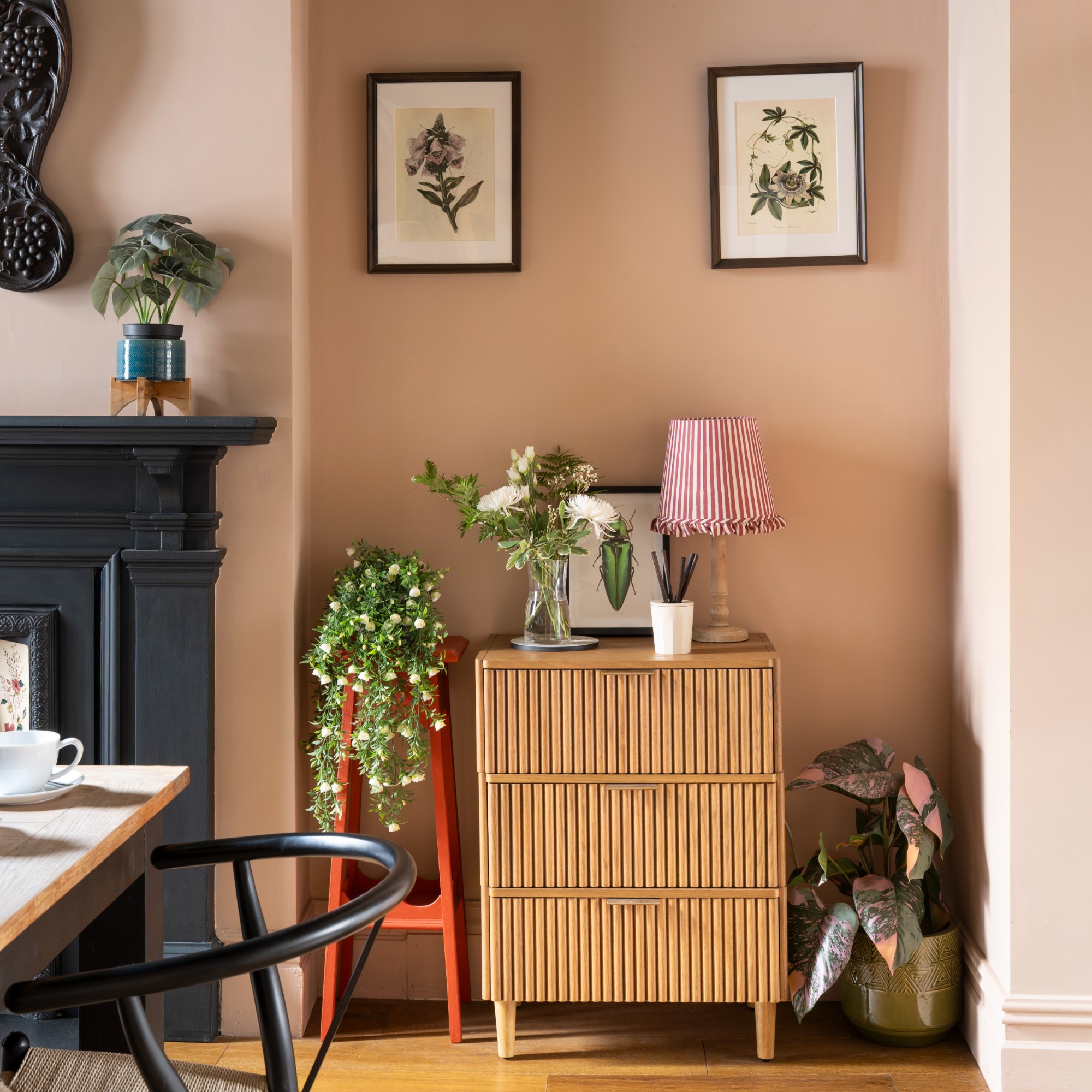 A pink dining room with a black fireplace and a chest of drawers displaying a vase of flowers and a table lamp with a striped shade