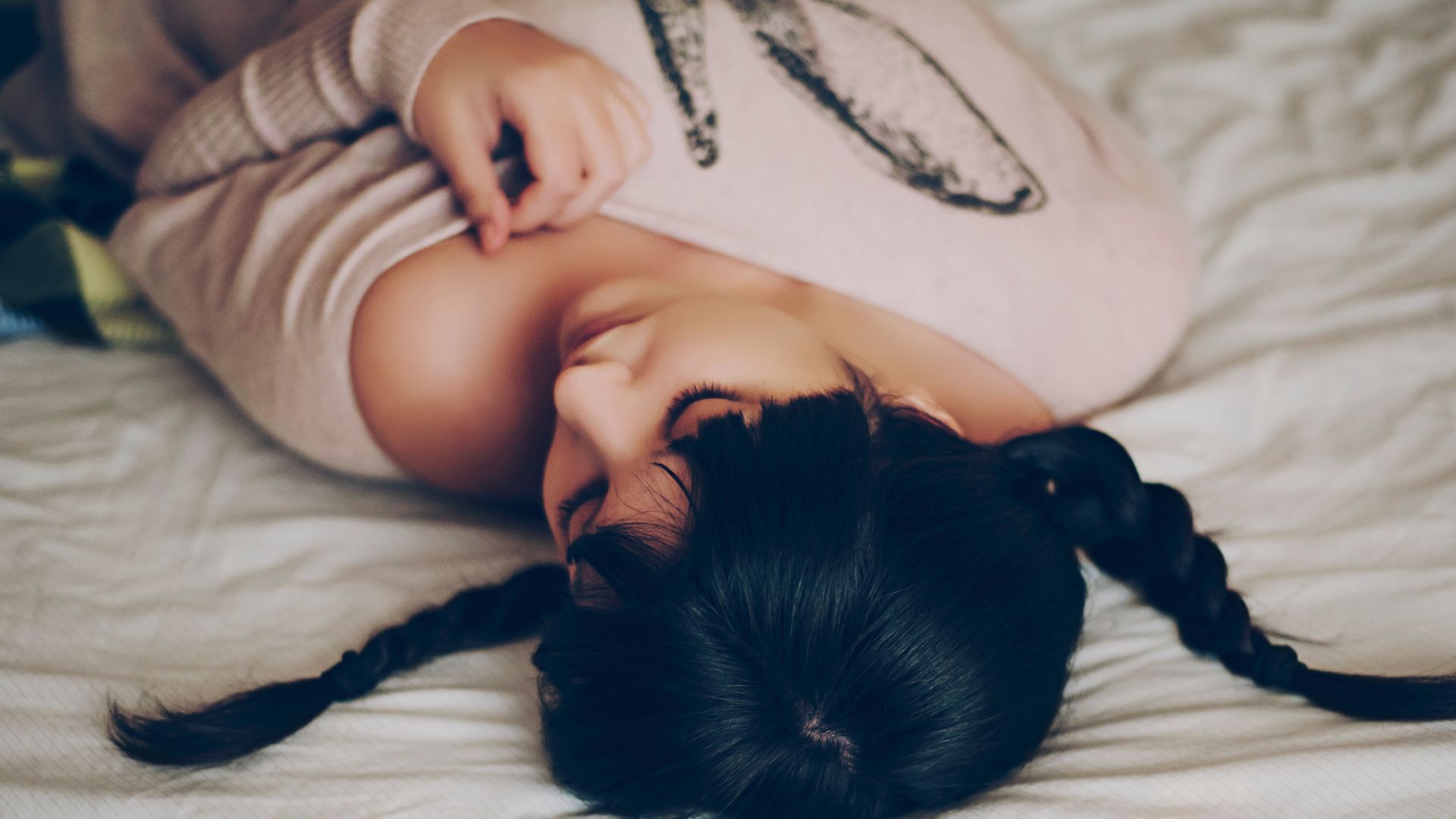 A woman with dark hair in plaits lies on her back in bed with her head turned to one side.