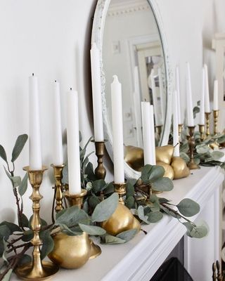 a collection of gold candlesticks and a eucalyptus garland on a white mantelpiece with round mirror