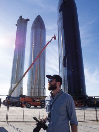 a bearded man stands holding a camera in front of some rockets
