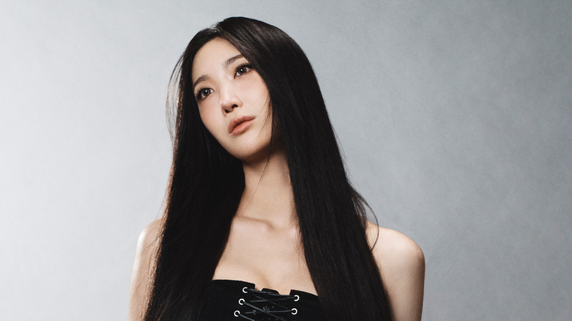 A portrait of singer-songwriter EJAE, wearing a lace-up corset and standing in front of a gray background.